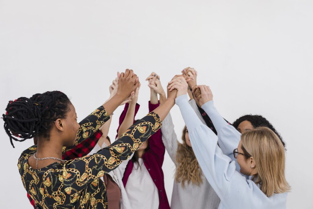 grupo de 6 mulheres em um círculo dando as mãos e eeguendo-as ao alto. A mulheres mais à esquerda tem pele negra e cabelos com dreadlock e está de costas