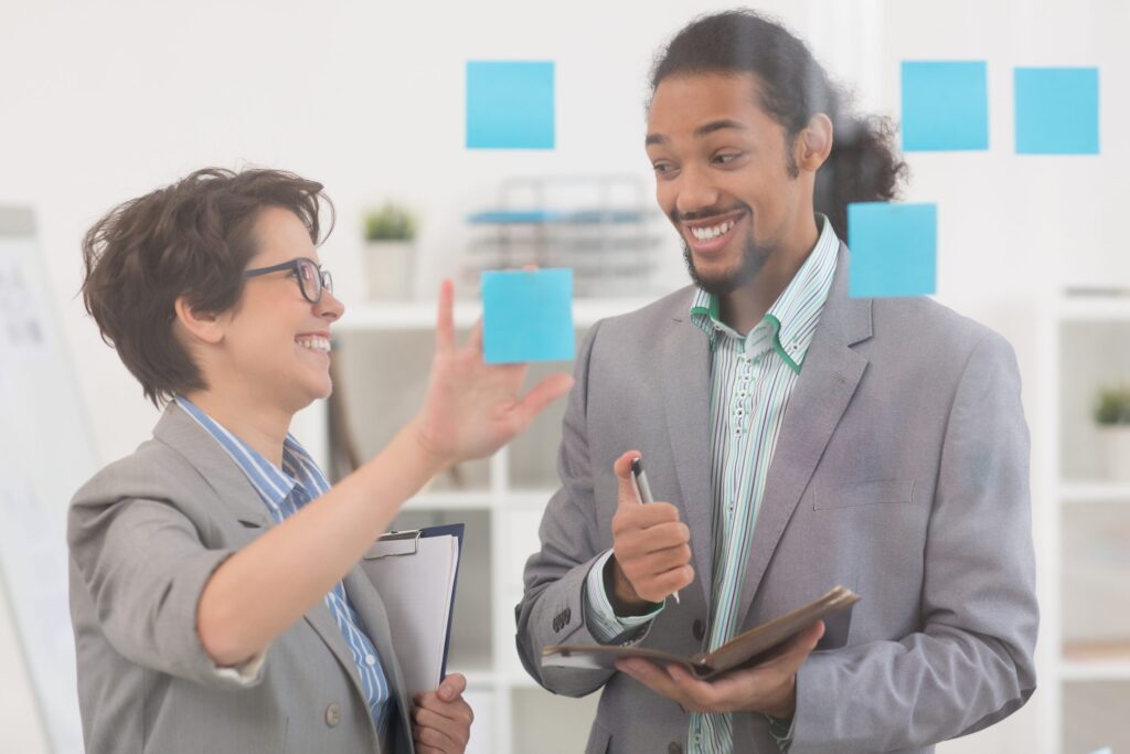 Imagem mostra ambiente de trabalho e dois colaboradores em um quadro fazendo conversando e fazendo planejamento. Eles pregam post its e expressam entusiasmo.|